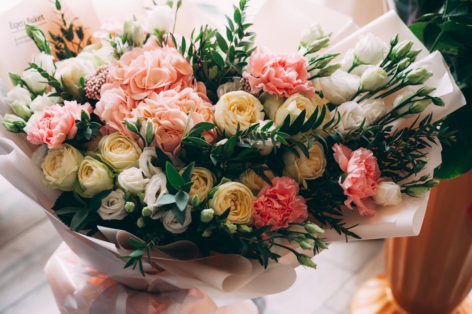 A bouquet of fresh flowers arranged with a mix of peach and white roses, white eustoma, and pink carnations, accented by green foliage with elongated leaves. The flowers are wrapped in elegant white t
