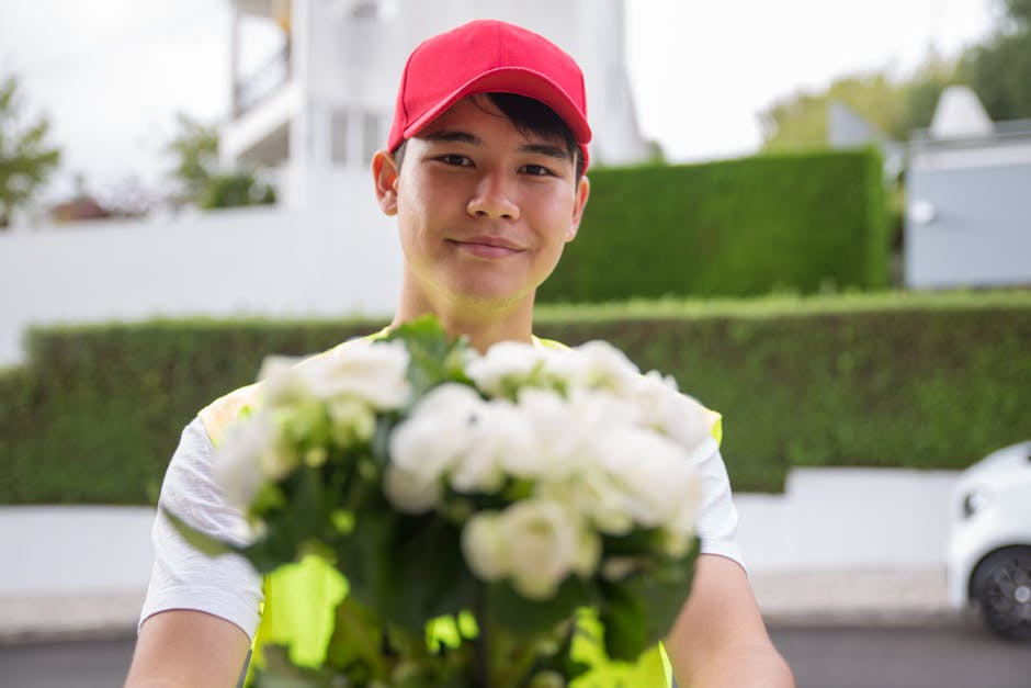 A delivery person in a red cap and white T-shirt holding a bouquet of fresh white flowers with green foliage, extending the floral arrangement towards the camera. The bouquet is wrapped in transparent
