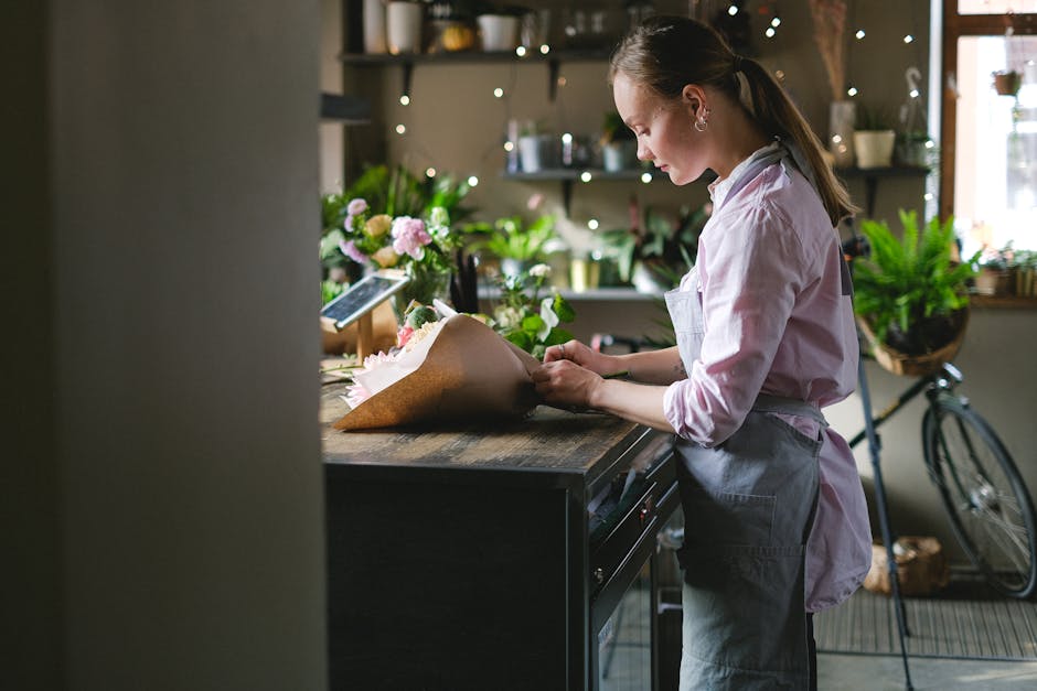 A female florist at a rustic wooden counter arranging a bouquet of fresh flowers wrapped in brown paper, with pink and white blooms visible, in a cozy flower shop decorated with green plants, fairy li