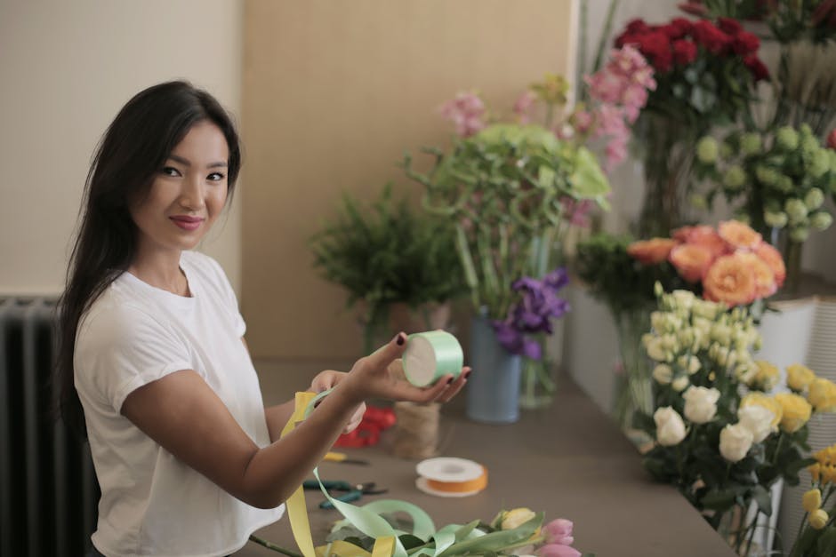 A young woman with long dark hair and a white T-shirt is smiling warmly while holding a roll of pastel green floral tape in her right hand and a piece of yellow wrapping paper in her left hand. She is