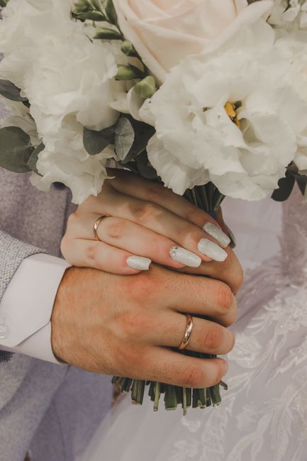 Close-up of a bride and groom's hands holding a bouquet of fresh white flowers, including lisianthus and glamelia, wrapped in dark green foliage. The bride's hand features glittery silver nail polish