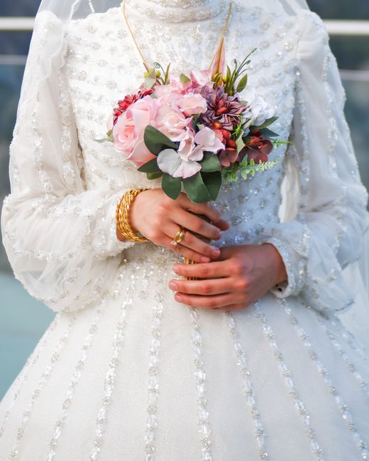 A bride wearing a long-sleeved, white wedding gown with intricate pearl embellishments and a high neckline, holding a vibrant floral bouquet. The bouquet features large pink roses, purple hydrangeas,