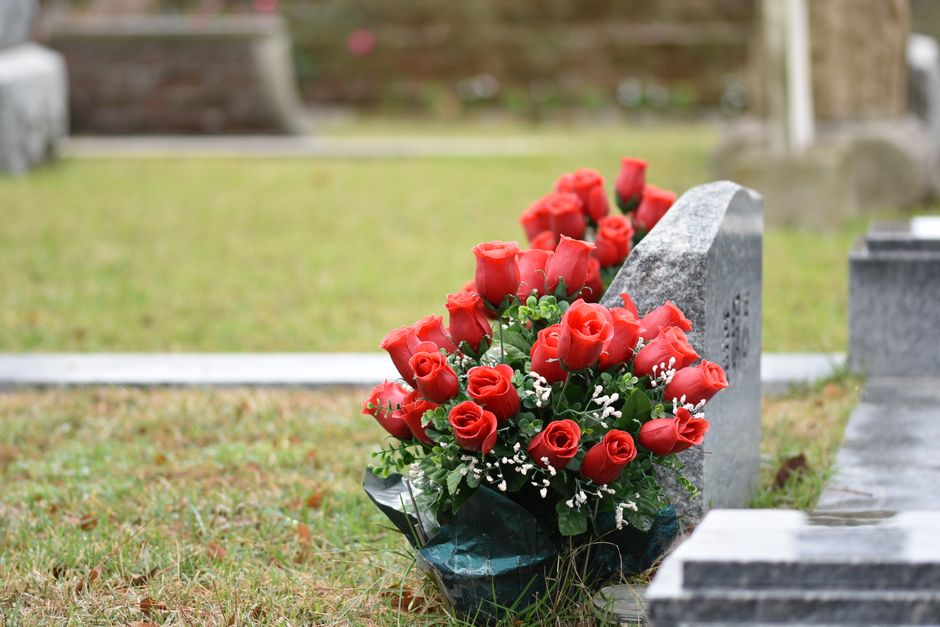 A bouquet arrangement of vibrant red roses with lush green foliage and small white accent flowers is placed at the base of a gray stone gravestone in a cemetery. The roses appear fresh with delicate,