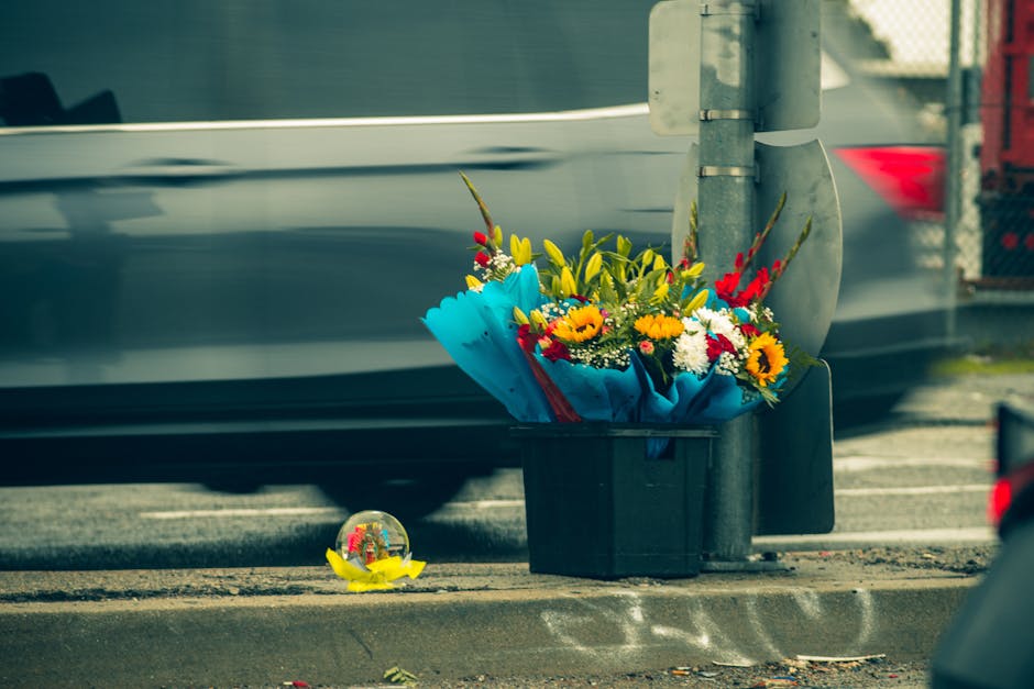 A floral arrangement of mixed fresh flowers, including sunflowers, red and white daisies, and yellow lilies, wrapped in blue paper and placed in a black container on the pavement beside a street pole.