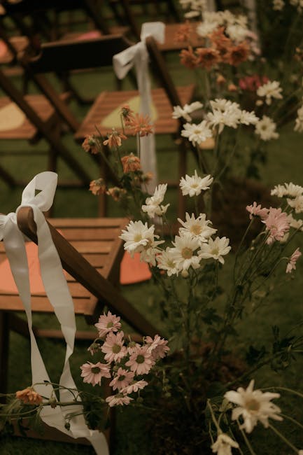 A floral arrangement featuring a mixture of white, pink, and peach daisies with delicate petals and green stems, placed outdoors among wooden chairs decorated with white ribbons. The daisies are in fu
